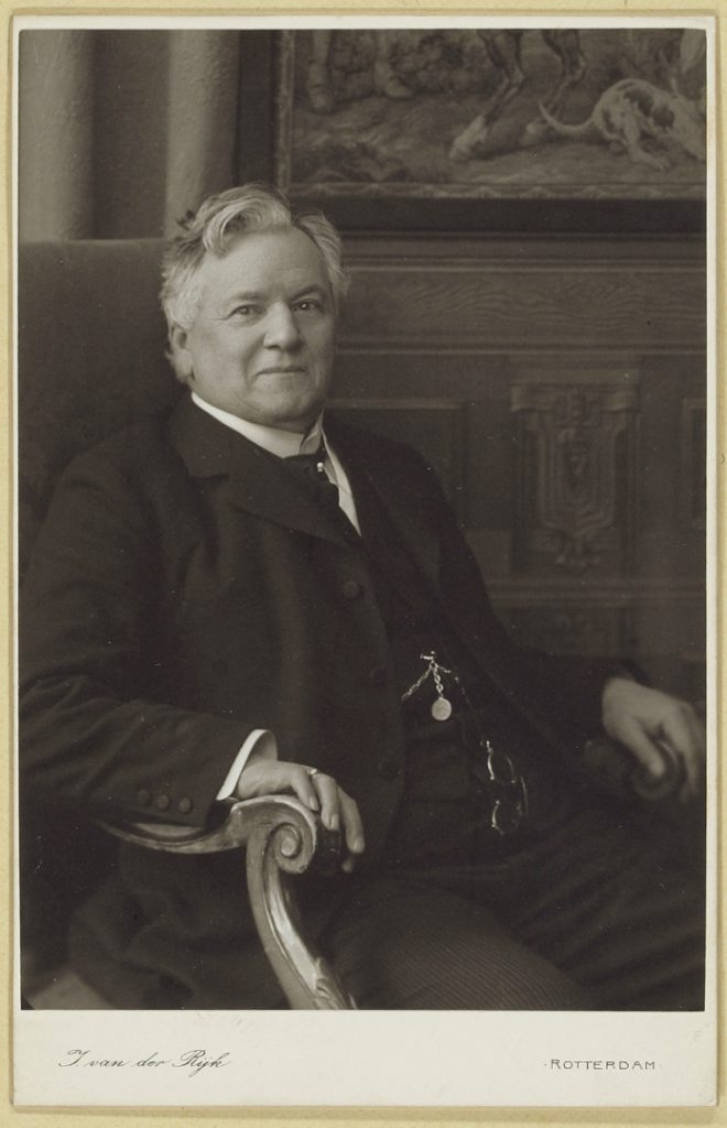 Black and white photograph of a middle aged male doctor sat in an ornate wooden chair
