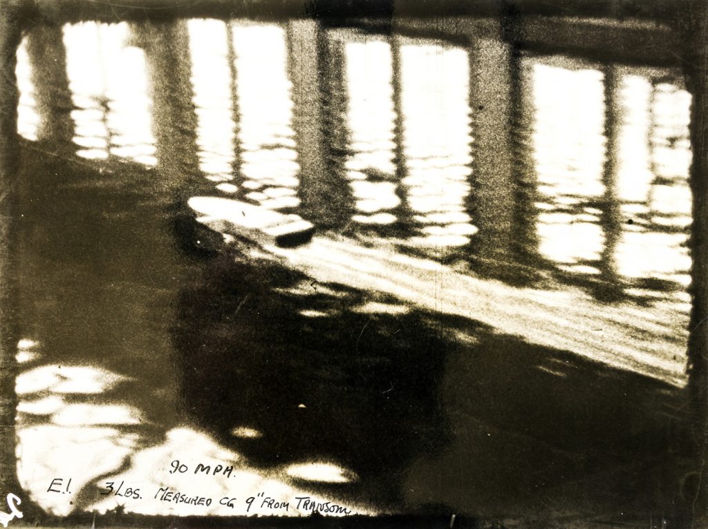 Black and white photograph of a model boat travelling at speed in a water tank testing facility
