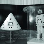 Black and white photograph of an exhibition display containing a spacewalk suit and a reentry module
