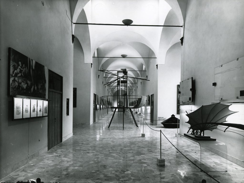 Black and white photograph of a corridor in which is displayed a number of leonardo models paintings and information plaques