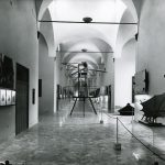 Black and white photograph of a corridor in which is displayed a number of leonardo models paintings and information plaques