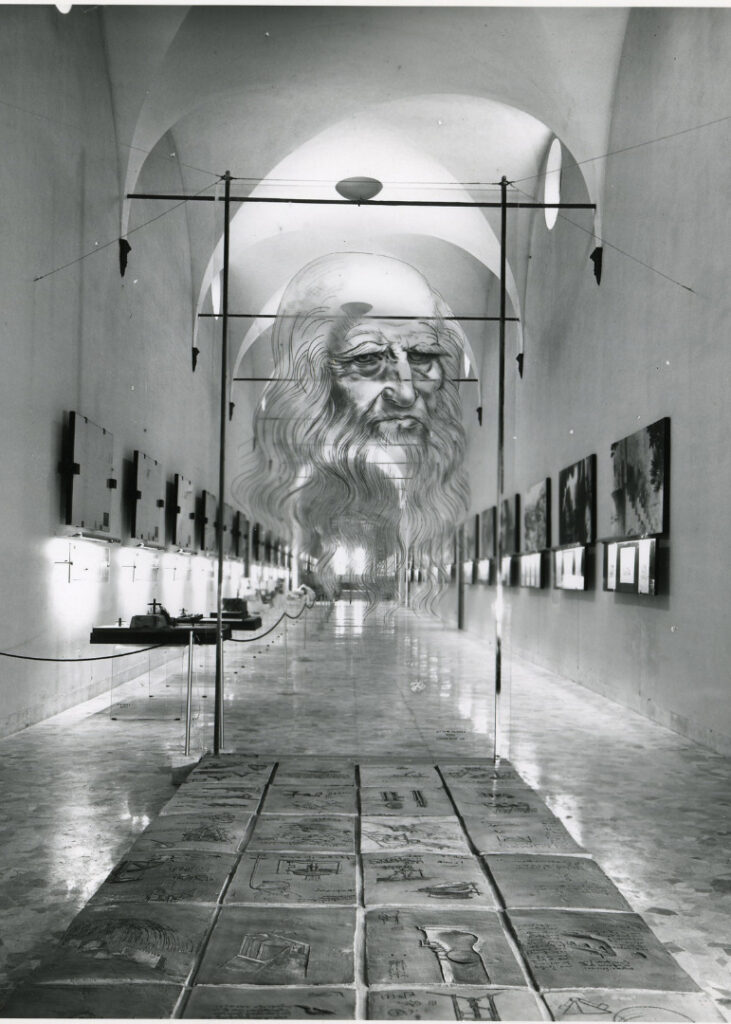 Black and white photograph of a corridor in which are displayed a number of leonardo models paintings and information plaques