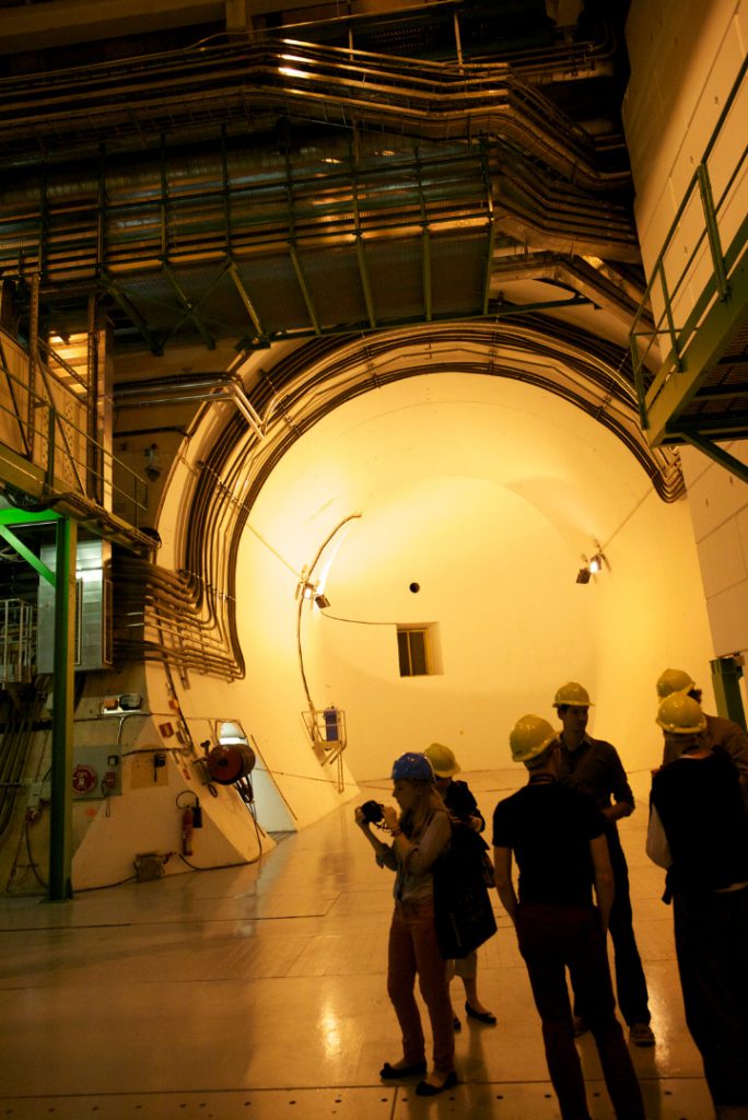Several visitors in hard hats stand in a large underground dimly lit room at the entrance to a large circular tunnel