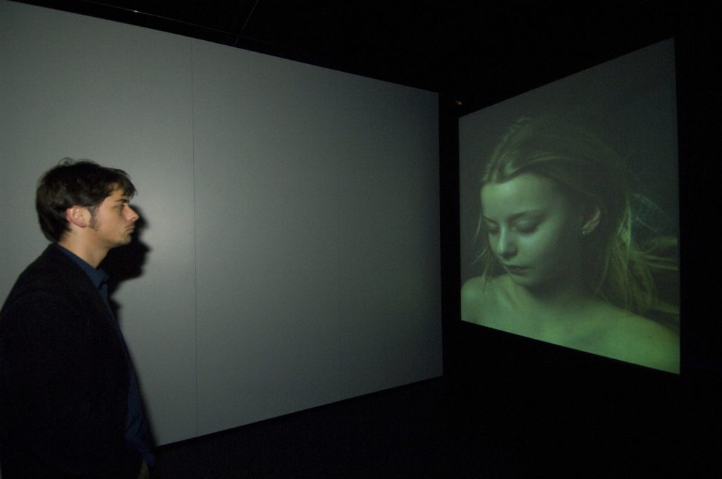 Colour photograph of a museum visitor looking at a video projection of a woman sleeping