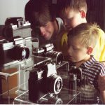 Colour photograph of a man and his two boys looking into a glass case display of cameras