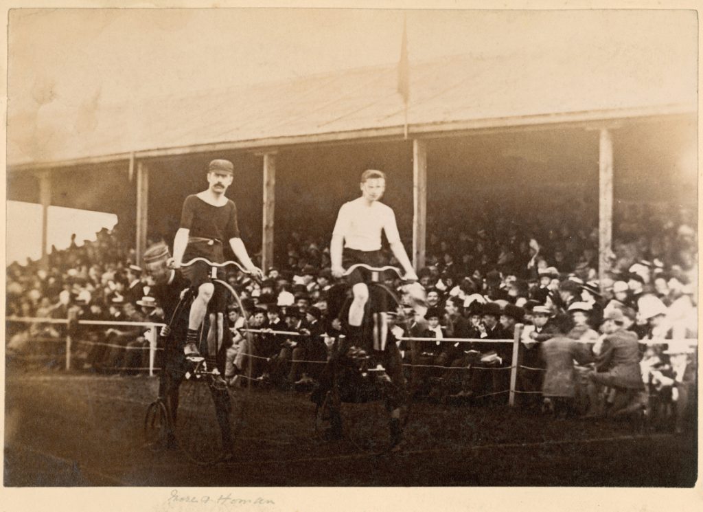 Sepia photograph of two cyclists on high wheeler bicycles at the start line of a race with spectators in the background from late nineteenth century