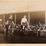 Sepia photograph of two cyclists on high wheeler bicycles at the start line of a race with spectators in the background from late nineteenth century