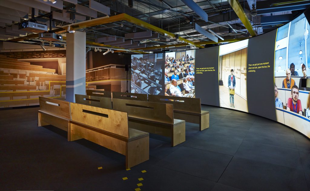 Part of the Collider exhibition showing several wooden benches in front of a large television screen showing the exhibition introductory video