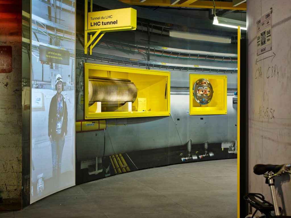 A recreation of the LHC tunnel in the Collider exhibition with LHC objects on display and a video projected onto a wall showing a CERN engineer giving information