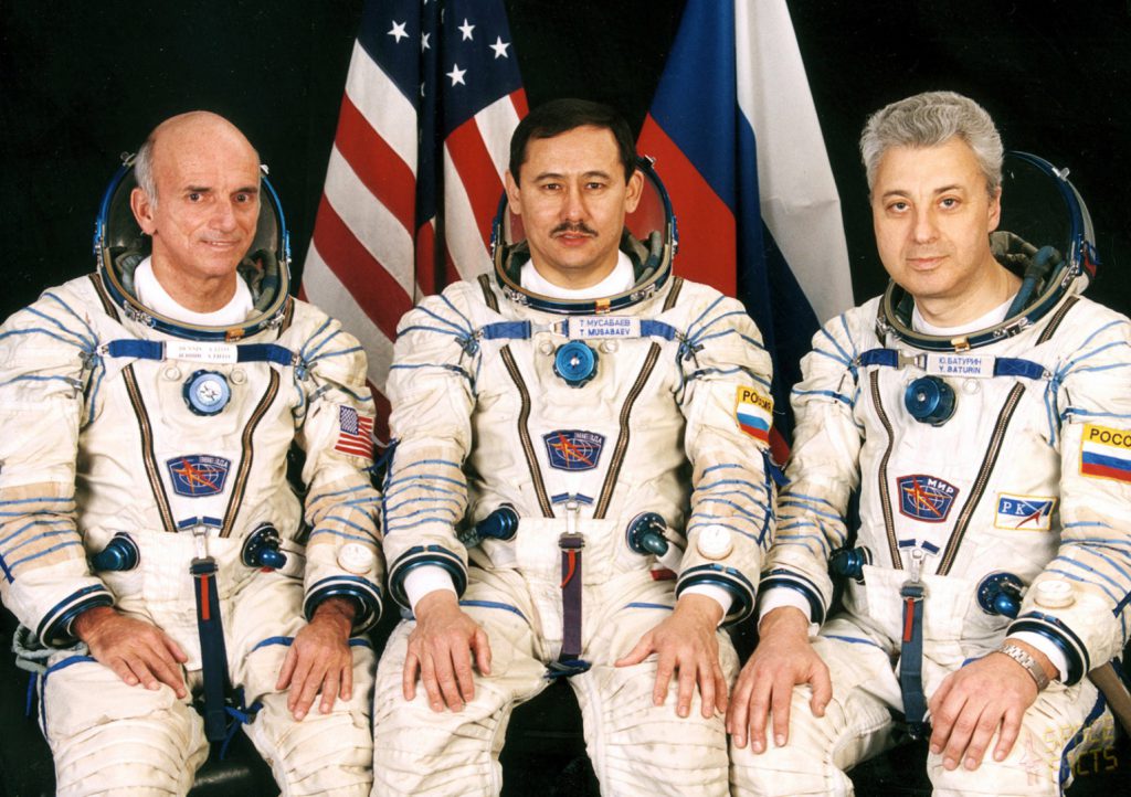 Colour photograph of three seated astronauts in space suits one of which is Dennis Tito the first space tourist
