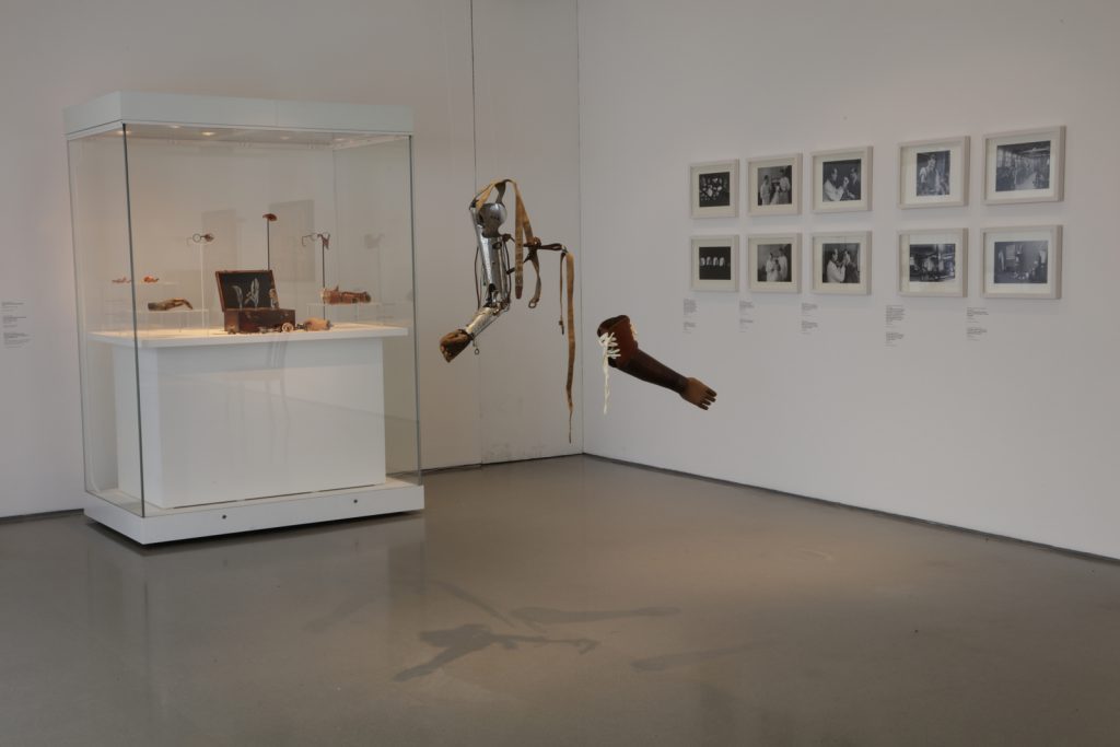 Colour photograph of an exhibition space displaying prosthetic arms hanging by wire