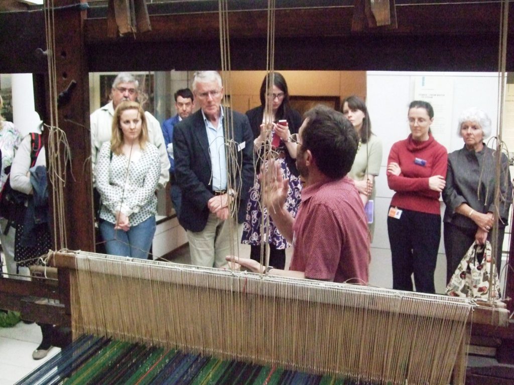 Colour photograph of of a weaver demonstrating how to work a loom in front of a museum audience