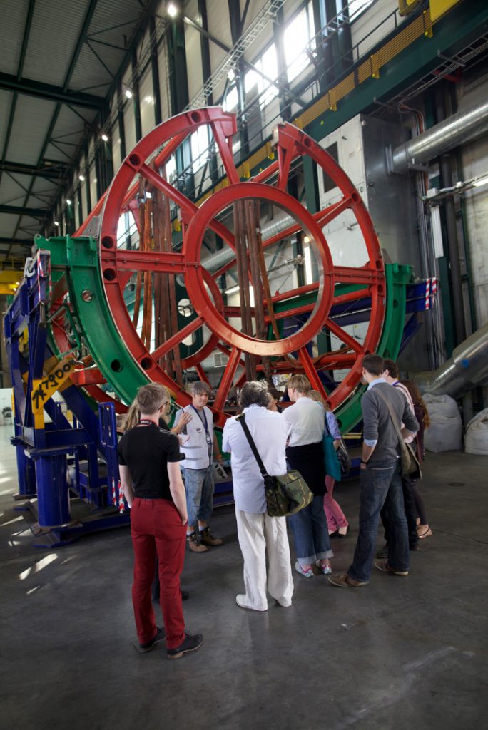 Several visitors listen to a CERN staff member talk in front of a large circular metal girder construction