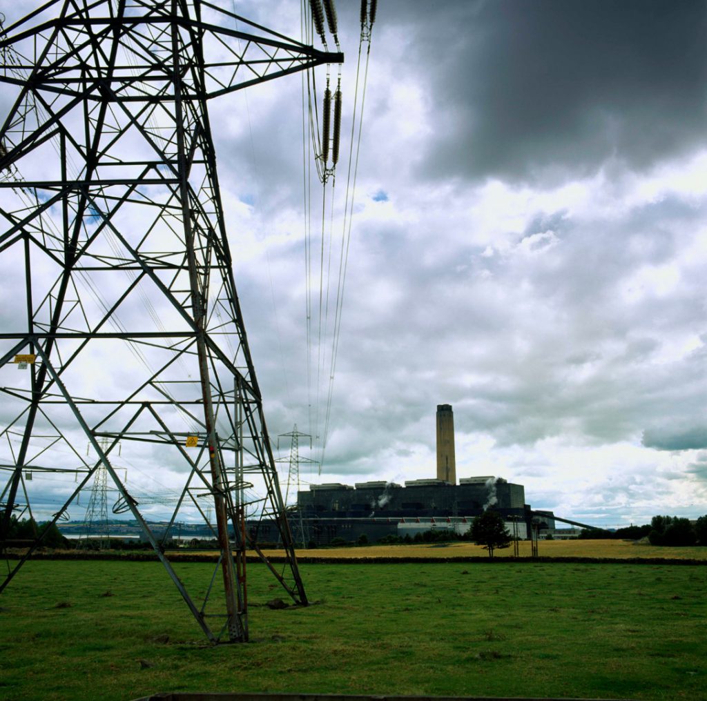 Colour photograph of an electricity pylon with a large energy facility in the background surrounded by fields and a lake
