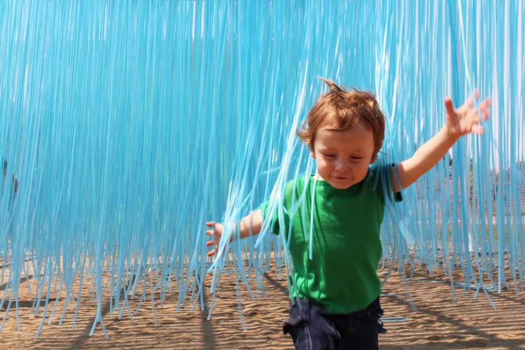 A toddler runs through a suspended collection of blue plastic strings