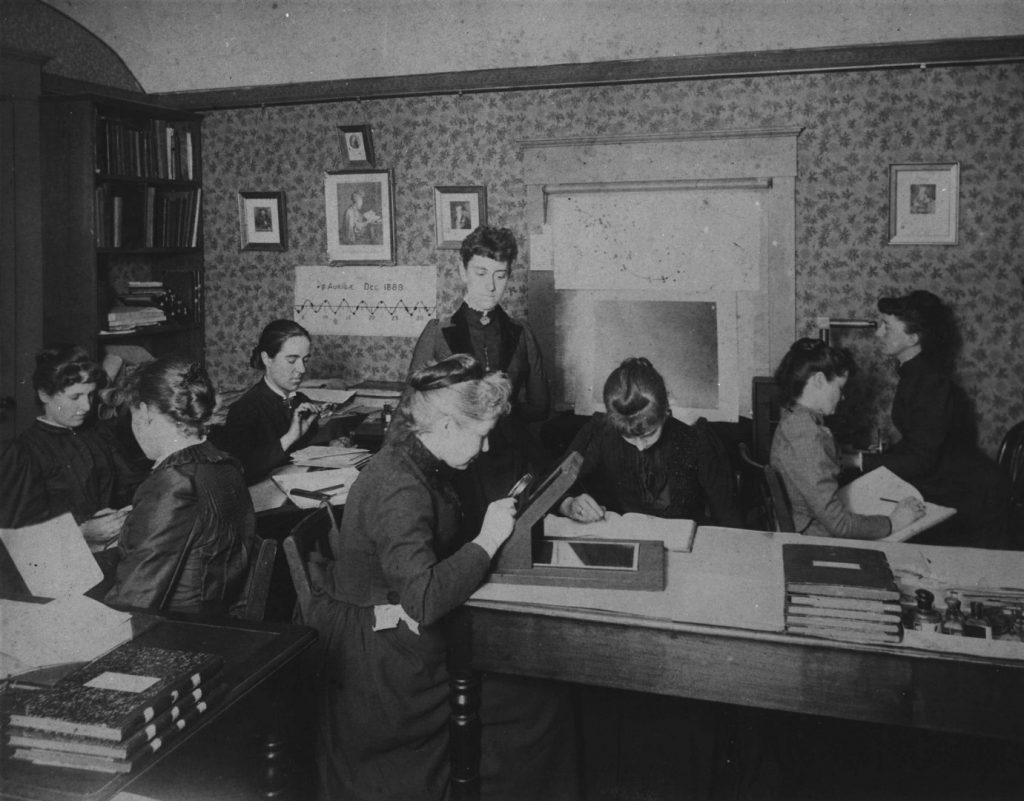 Black and white photograph from the 1880s of 8 women attempting to classify stars from recorded observations