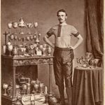 Sepia photograph of a cyclist with a case of trophies from the late nineteenth century