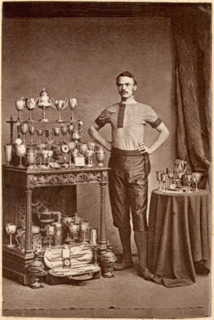 Sepia photograph of a cyclist with a case of trophies from the late nineteenth century