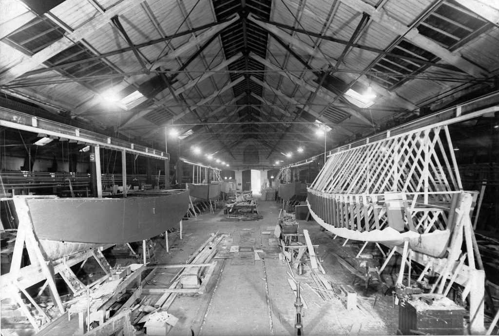 Black and white photograph of a large warehouse with two partially constructed boats