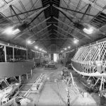 Black and white photograph of a large warehouse with two partially constructed boats