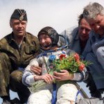 Colour photograph of Helen Sharman female space tourist in a space suit smiling and holding flowers as she is helped down from a module