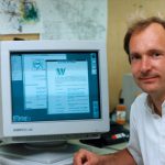 Photograph of Tim Berners-Lee sitting next to a computer display on which the words world wide web initiative are legible as well as an old CERN logo