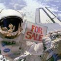 Colour photograph of an astronaut in space floating above a spacecraft and holding up a sign which reads for sale