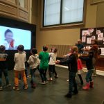 Colour photograph of children copying a dance from a screen in a museum gallery