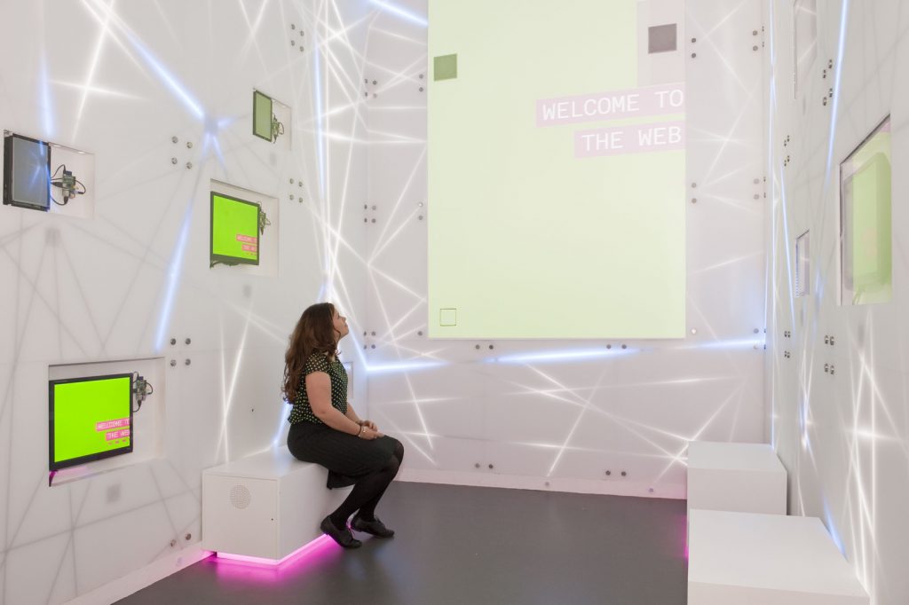 Colour photograph of the inside of a colourful and brightly lit installation in the Information Age exhibition showing a woman looking at a large screen with the text welcome to the web