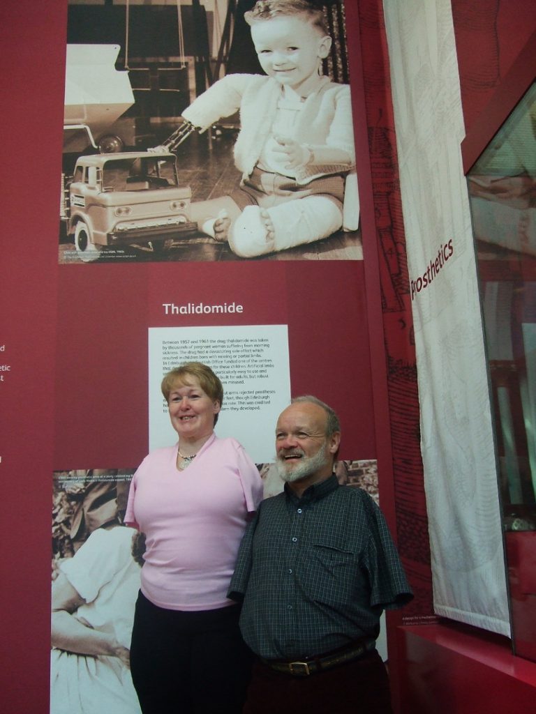 Colour photograph of a male and a female thalidomide patient standing in front of an exhibition display wall
