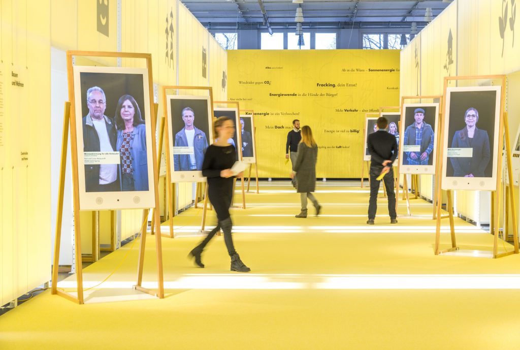 Colour photograph of visitors inside a large gallery space with photographs displayed on easels