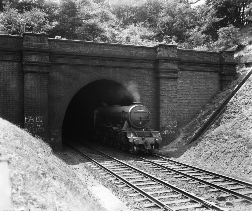 Black and white photograph of the Flying Scotsman steam train emerging from a tunnel. Anti diesel train graffiti is visible on the brickwork
