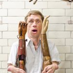 Colour photograph of Mat Fraser holding up two historical wooden prosthetic arms