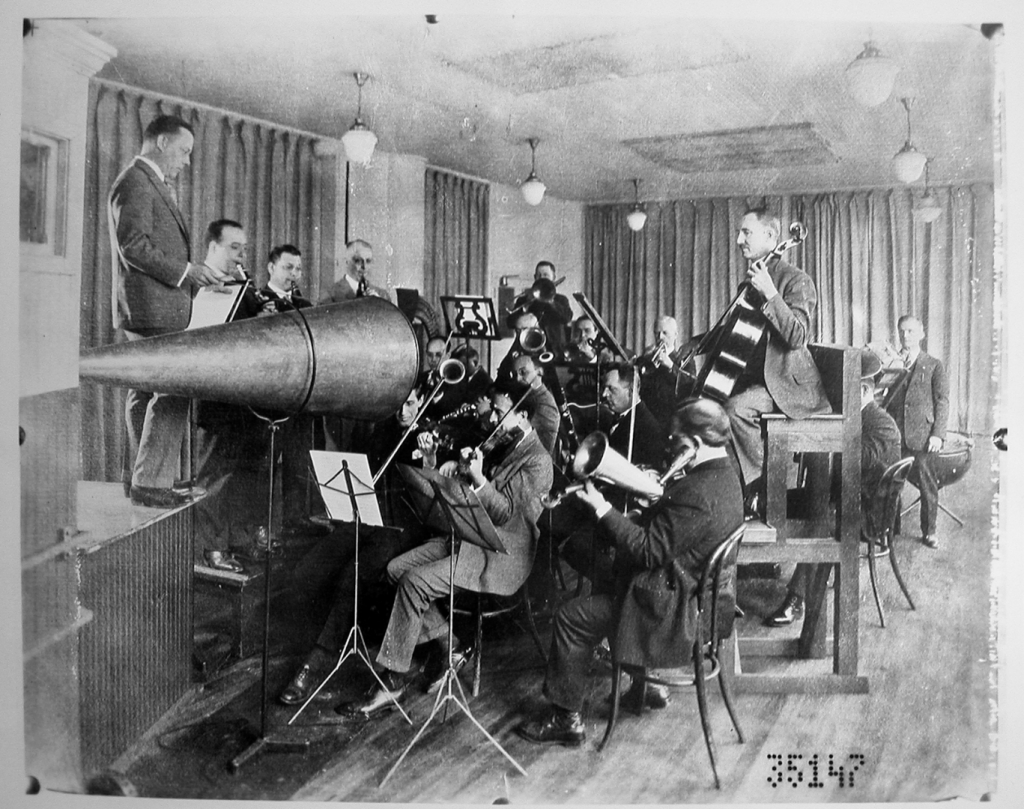 Black and white photograph of a studio orchestra playing into a megaphone shaped recording instrument