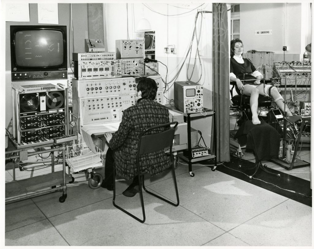 Black and white photograph of a neurologist seated before an EEG with a patient in the adjacent room