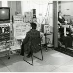 Black and white photograph of a neurologist seated before an EEG with a patient in the adjacent room