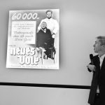 Black and white photograph of Mat Fraser alongside an old German poster depicting a doctor and a disabled character