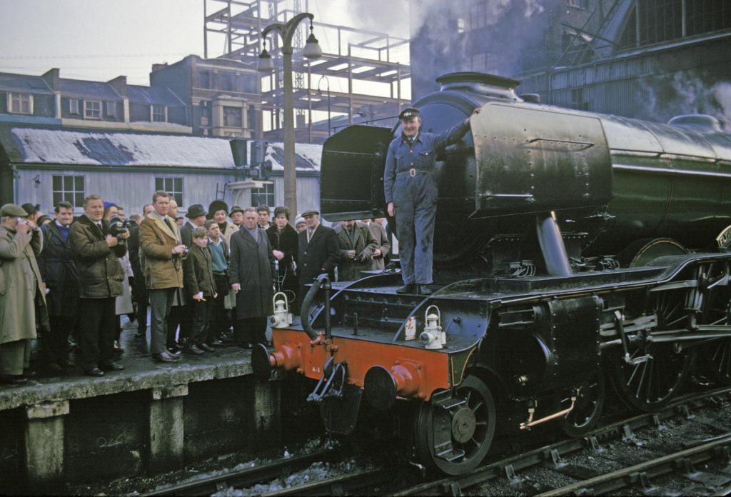 Colour photograph of the Flying Scotsman steam train on track at London Kings Cross station with the restorer Alan Spegler standing on the front