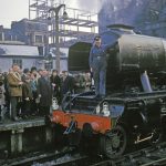Colour photograph of the Flying Scotsman steam train on track at London Kings Cross station with the restorer Alan Spegler standing on the front