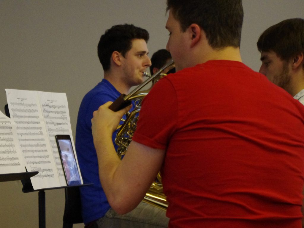 Colour photograph of a member of RCM playing the french horn with the conducting being done via a tablet computer