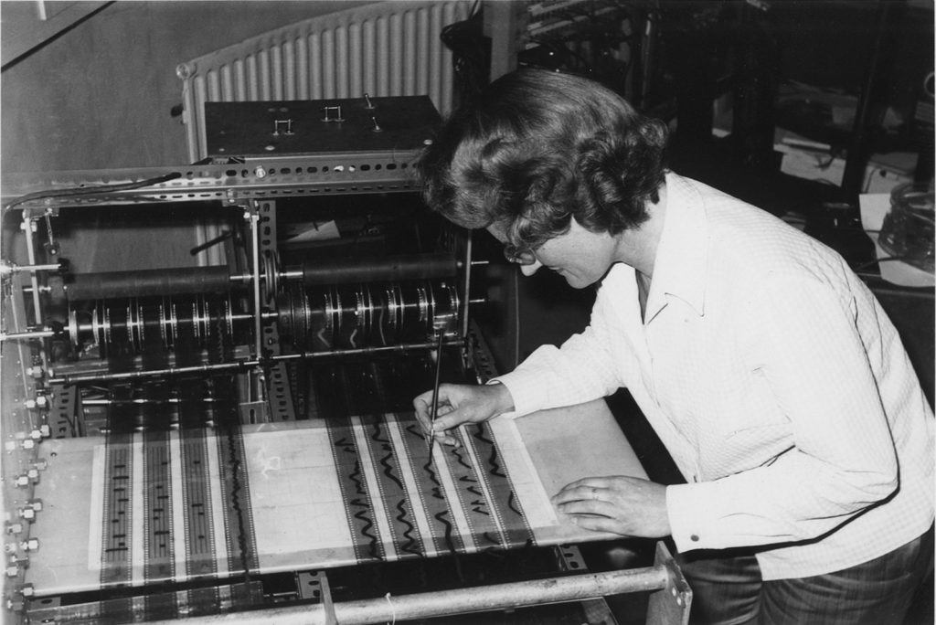 Black and white photograph of Daphne Oram leaning over the Oramics Machine and drawing black lines on one of the acetate strips