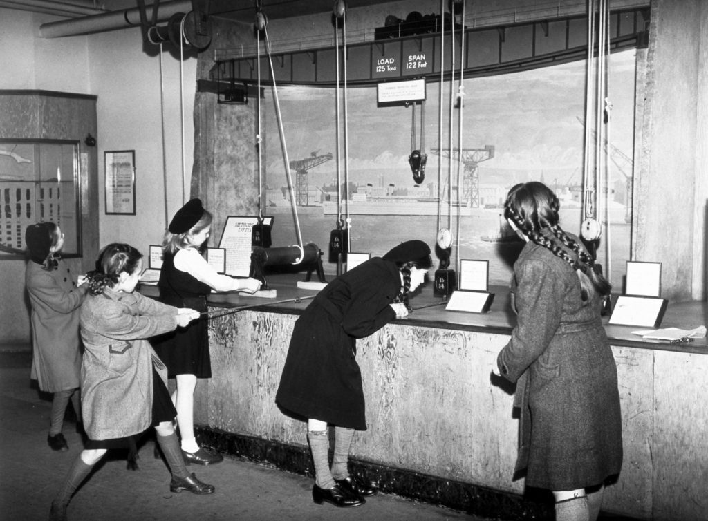 Black and white photograph of schoolgirls lifting weights using pulleys at a museum exhibition