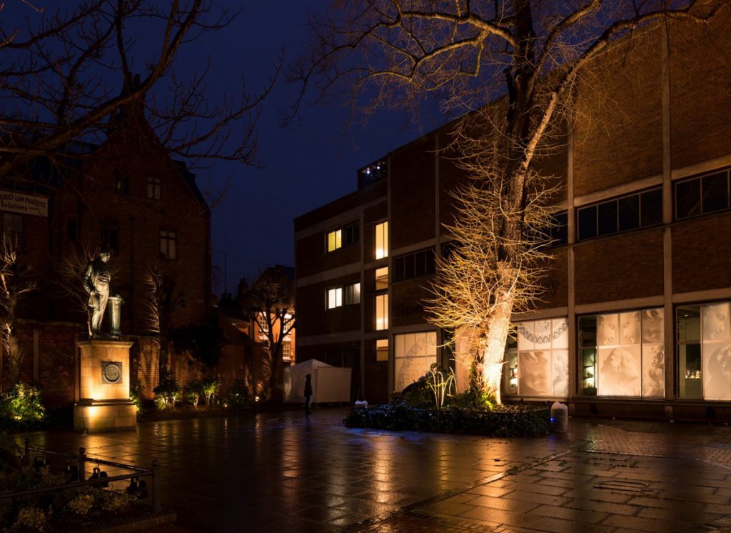 Colour photograph of a building and courtyard with lighting at the base of a tree and a statue