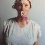 Colour photograph of Jo Spence dressed in baby clothes with dummy