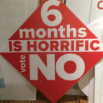 A large protest sign reading 6 months is horrific vote no