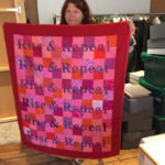 A woman holds up a blanket stitched with the words Rise and Repeal