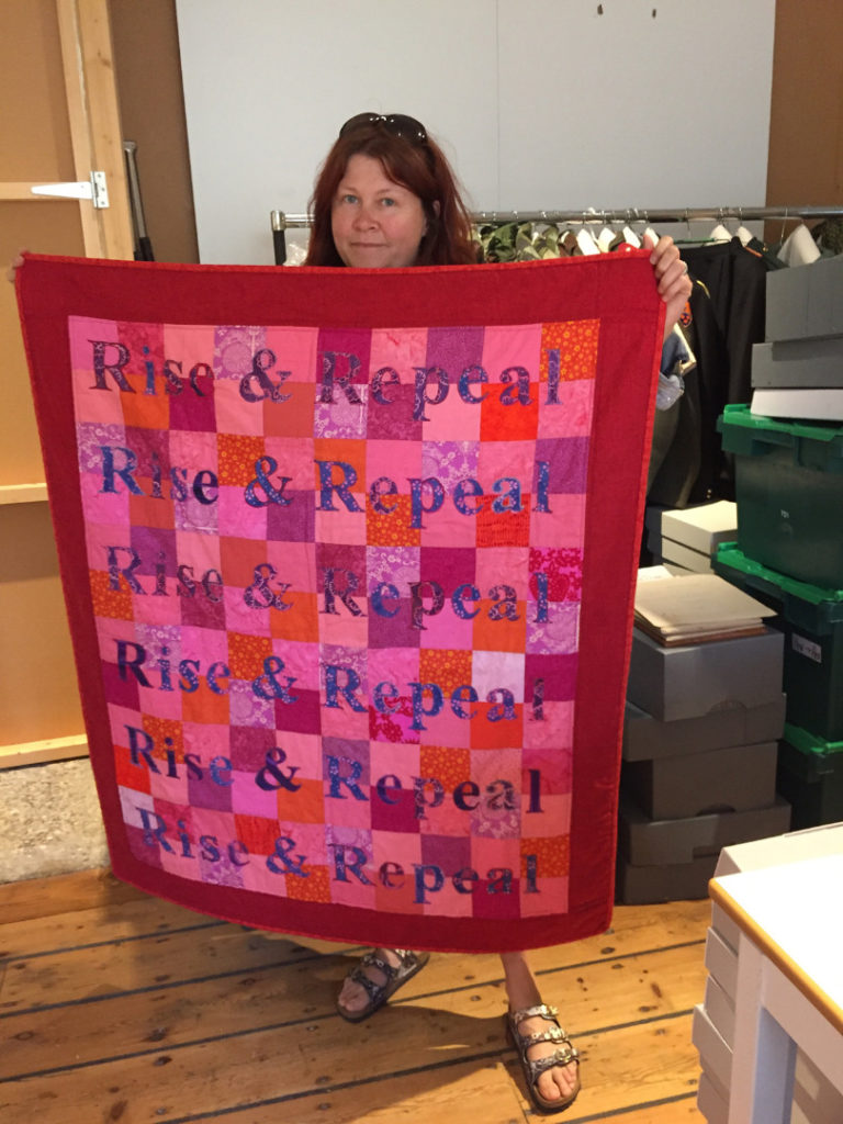 A woman holds up a blanket stitched with the words Rise and Repeal