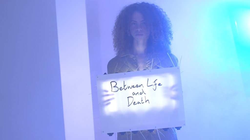 Video still showing a woman holding a sign saying Between Life and Death