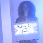 Video still showing a woman holding a sign saying Between Life and Death