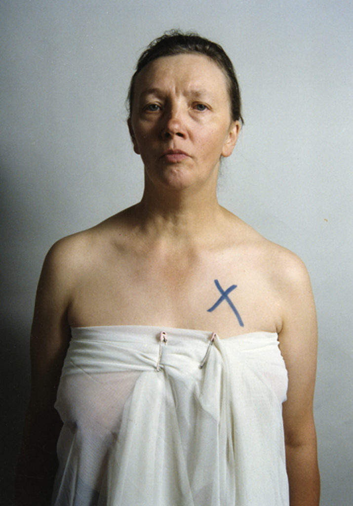Colour photograph of Jo Spence wrapped in a sheet with a black cross over her breast